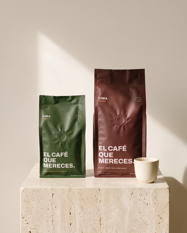 El de Diario: bolsa vino de CIMA Origen de 500 g junto a una bolsa verde de CIMA Reserva de 250 g, sobre un cubo de travertino con una taza de café