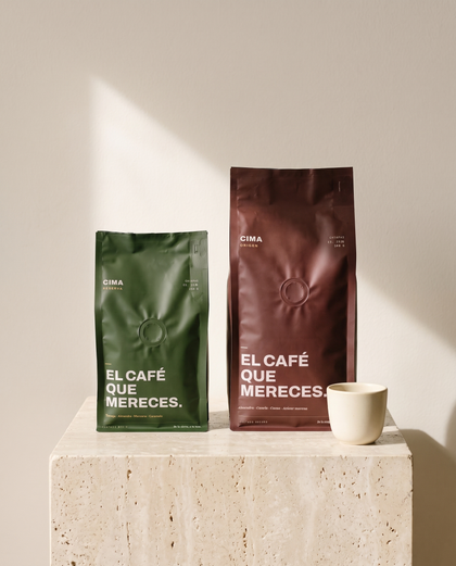 El de Diario: bolsa vino de CIMA Origen de 500 g junto a una bolsa verde de CIMA Reserva de 250 g, sobre un cubo de travertino con una taza de café