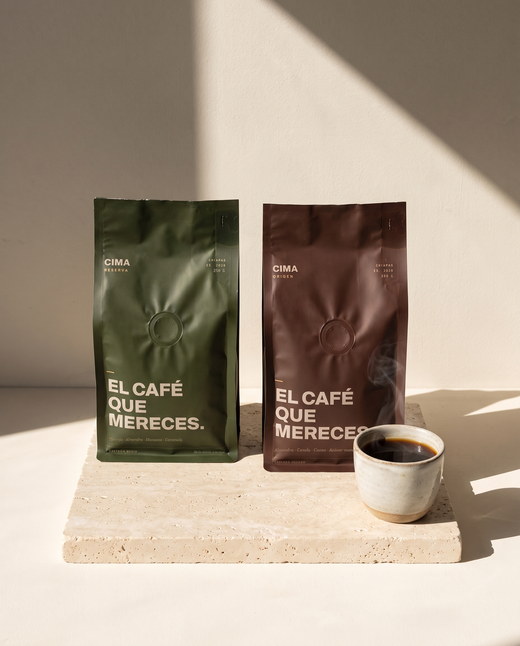 El Dúo: bolsa verde de CIMA Reserva y bolsa vino de CIMA Origen, 250 g cada una, sobre una placa de travertino con una taza de café al lado