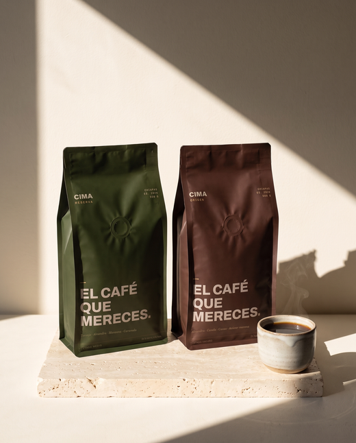 El Kilo: bolsa verde de CIMA Reserva y bolsa vino de CIMA Origen, 500 g cada una, sobre una losa de travertino con una taza de café al lado